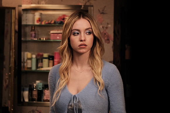 A blonde woman with blue eyeshadow and a blue cardigan looks thoughtfully to the side.