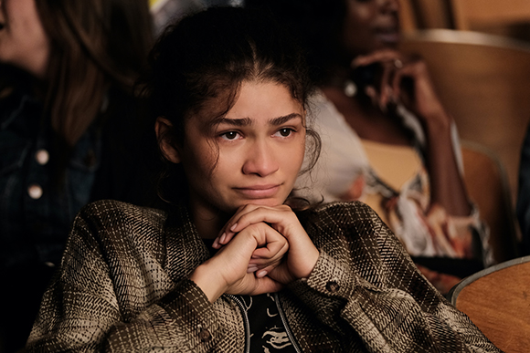 A young woman with dark hair looks forward with a sad, pained expression, hands clasped under her chin.