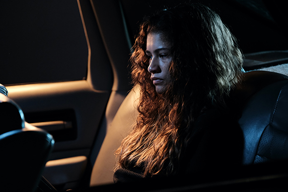 A young woman with long, curly hair sits in a dark car, illuminated by dim light.