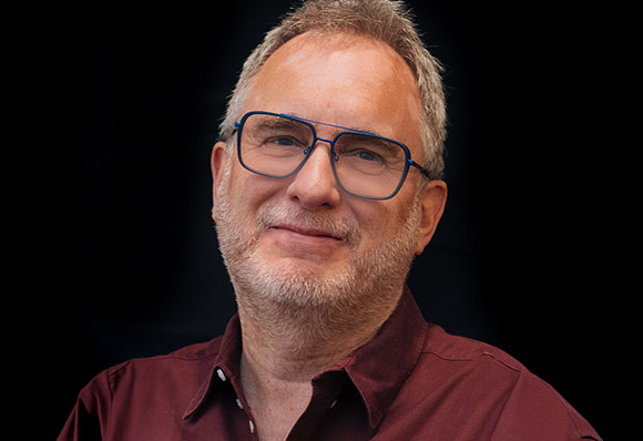 A smiling man with graying hair and a beard wearing blue-rimmed glasses and a maroon shirt.