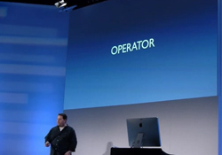 A man stands on a stage next to a computer monitor, facing a large blue screen that reads 'OPERATOR'.