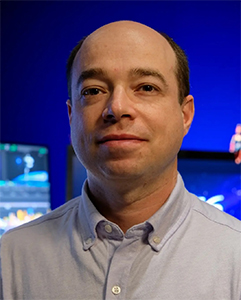 A man with a receding hairline wearing a light blue button-down shirt against a blue background.