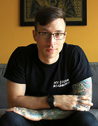A man with short dark hair and glasses sits on a gray couch, wearing a black t-shirt and showing arm tattoos.