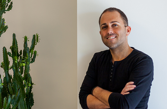 A smiling man in a black long-sleeve shirt stands with arms crossed next to a tall cactus.