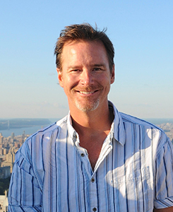 A smiling man with short brown hair wearing a blue and white striped button-down shirt.
