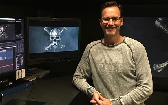 A man in a grey sweatshirt smiles in a dark room with video editing monitors.
