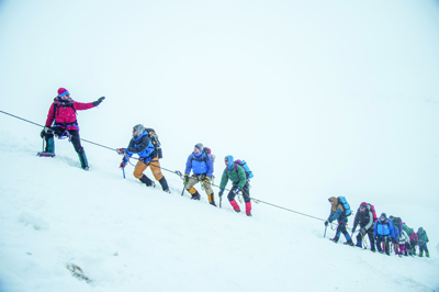 A line of climbers in colorful gear ascending a steep, snowy mountain slope using a guide rope.