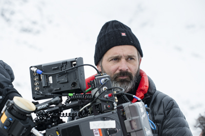 A bearded man in a black beanie and puffer jacket looking past a large professional film camera.