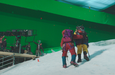 Actors in climbing gear walk on a snowy slope in front of a large green screen in a studio.