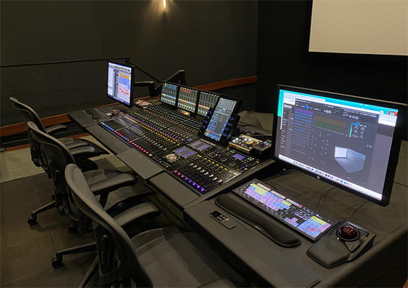 A large digital audio mixing console with multiple monitors and control surfaces in a dark studio.