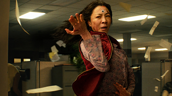 A woman with a third eye on her forehead reaches forward in a chaotic office with flying papers.