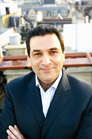 A man in a dark suit and light blue shirt smiling with a city rooftop background.