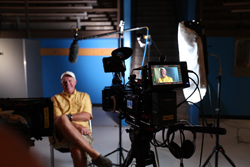 A man in a yellow shirt sits for an interview in front of a professional video camera and lighting equipment.