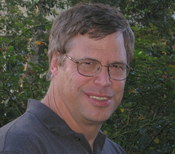 A man with short brown hair and glasses smiling outdoors against a background of green leaves.
