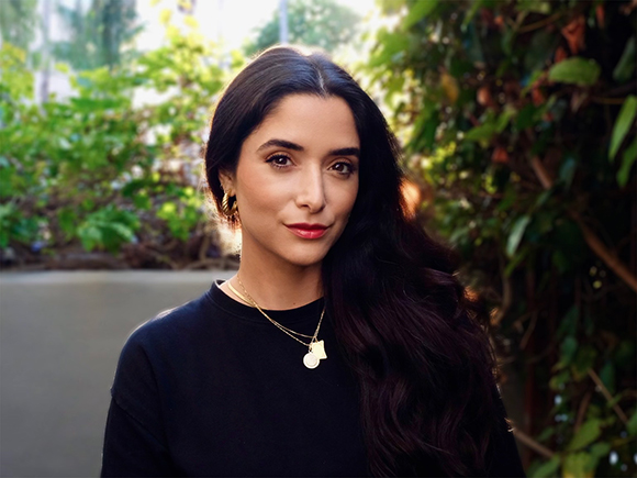 A woman with long dark hair and gold earrings wearing a black top, standing in front of green foliage.