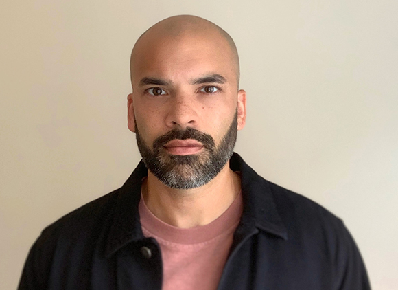 A bald man with a dark beard and mustache wearing a dark jacket over a pink shirt.