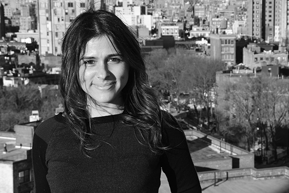 A smiling woman with long dark hair posing outdoors with a cityscape in the background.