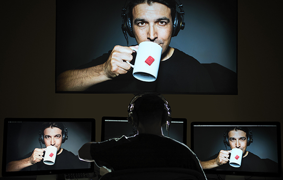 A person in headphones watches a large screen showing a man drinking from a white mug with a red logo.