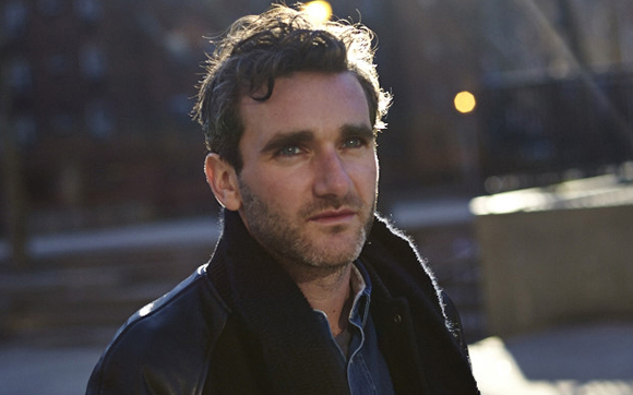 A man with wavy dark hair and stubble looks into the distance outdoors during golden hour.