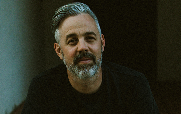 A man with graying hair and a beard wearing a black shirt looks toward the camera.