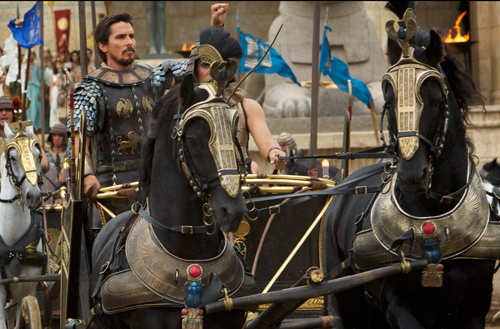 A man in ornate silver and black armor stands behind a chariot pulled by two black horses.