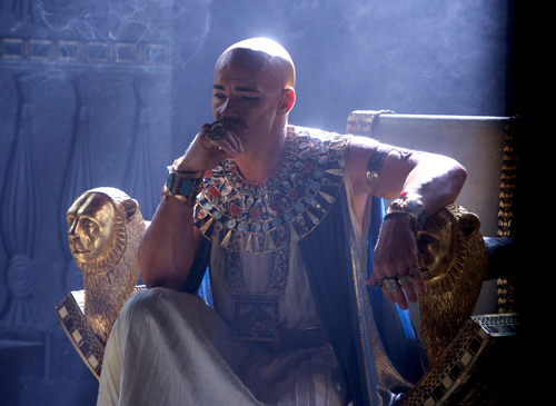 A bald man in elaborate gold and blue Egyptian-style clothing sits on a golden lion-headed throne.