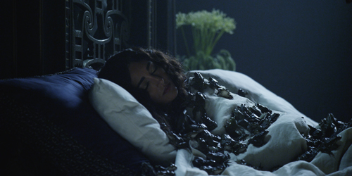 A person lies in a bed with white sheets, covered in numerous dark oysters.