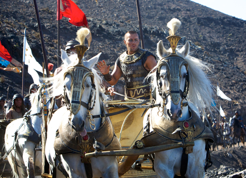 A man in ornate black and gold armor stands in a chariot pulled by two white horses.