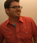 A smiling man with glasses wearing a red and white checkered short-sleeved button-down shirt.