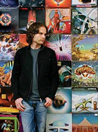 A man with long hair wearing a black jacket and blue jeans stands before a wall of colorful record covers.