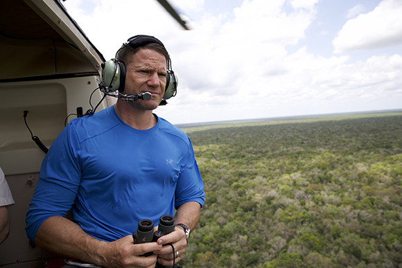 A man wearing headphones and a blue shirt looks out from an open helicopter door over a vast green forest.