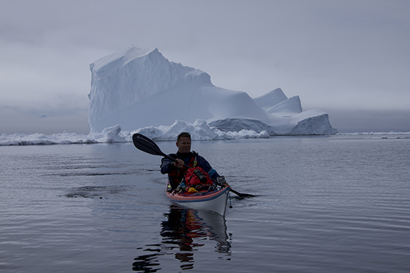 A person in a red and white kayak paddles across calm water toward a massive iceberg under a gray sky.