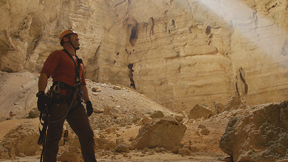A climber in a red shirt and safety gear stands inside a large, sunlit rocky cavern.