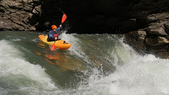 A person in an orange helmet and blue jacket paddles an orange kayak through turbulent river rapids.