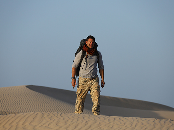 A man with a large backpack walks across rippled sand dunes under a clear sky.