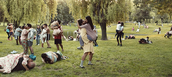 Various people embracing, hugging, or lying on the grass in a lush green park near a pond.