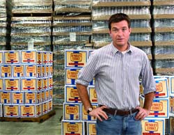 A man in a striped shirt and jeans stands in a warehouse surrounded by stacks of boxes.