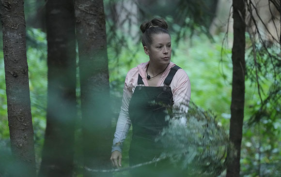 A woman wearing pink long-sleeved shirt and dark overalls walks through a dense, green forest.