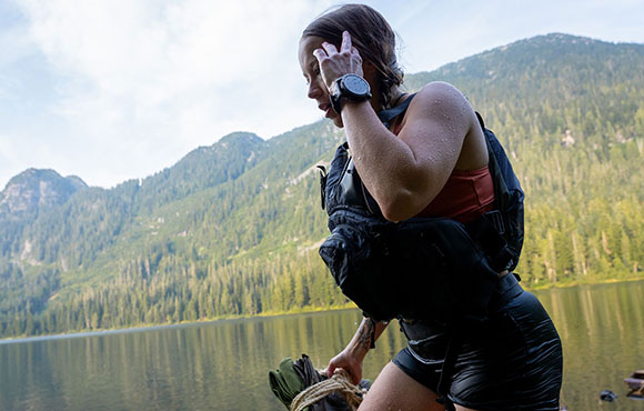 A woman in a black life vest stands by a calm lake with forested mountains behind her.