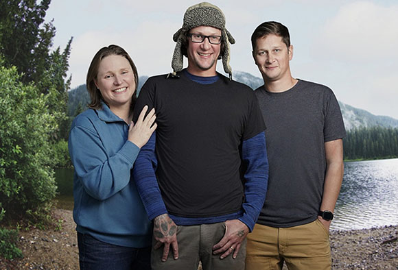 A woman in blue and two men, one in a black shirt and hat, standing by a lake with mountains in the background.