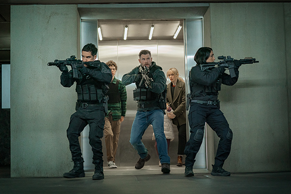 Three tactical officers with rifles flank two civilians inside a brightly lit elevator.
