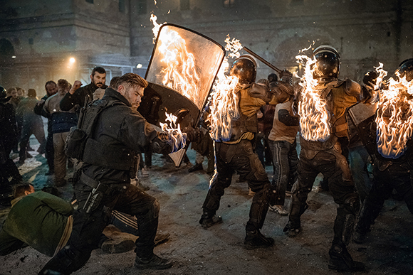 Riot police in helmets and armor face protesters amidst bright flames and thick smoke at night.