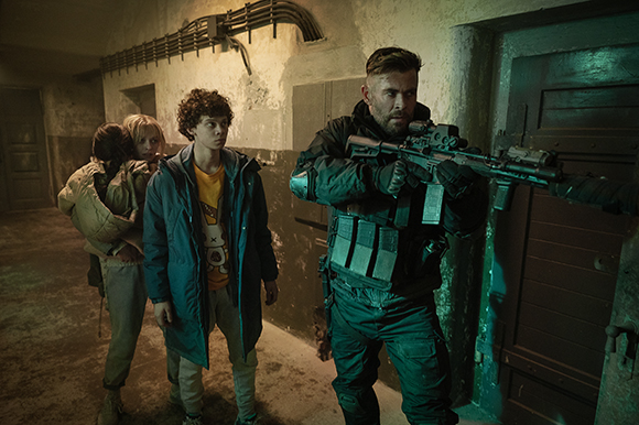 A man in dark tactical gear aims a rifle in a dim hallway near two young people.