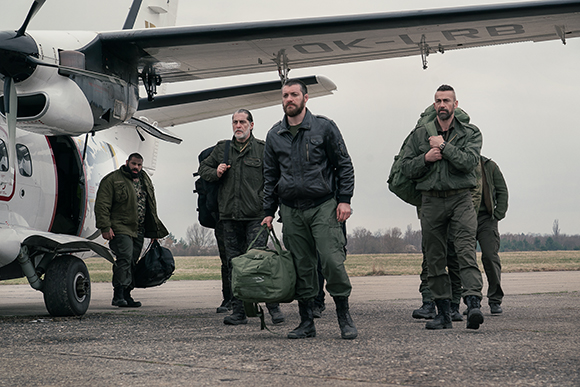 Four men in olive green and dark clothing stand on an airfield next to a white propeller plane.