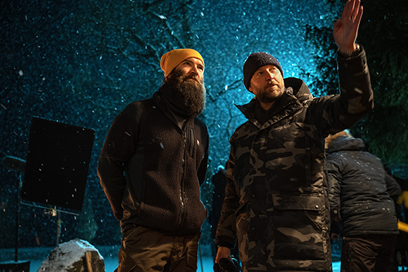 Two bearded men in winter gear stand outside at night as snow falls around them.