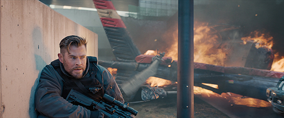 A man in a dark jacket holds a rifle while hiding behind a concrete wall near a burning helicopter.
