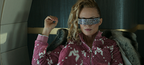 A woman with blonde curly hair wearing a pink quilted jacket and sparkling, jeweled sunglasses.