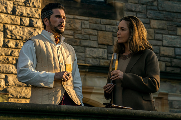 A man in a white vest and a woman in a dark blazer hold champagne glasses while looking at each other.