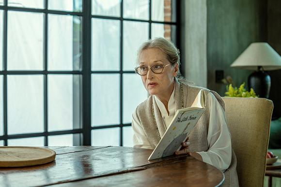 An older woman with glasses sits at a wooden table, looking up while holding an open book.