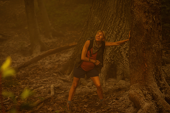 A woman in hiking gear leans against a thick tree trunk in a dusty, sunlit forest.